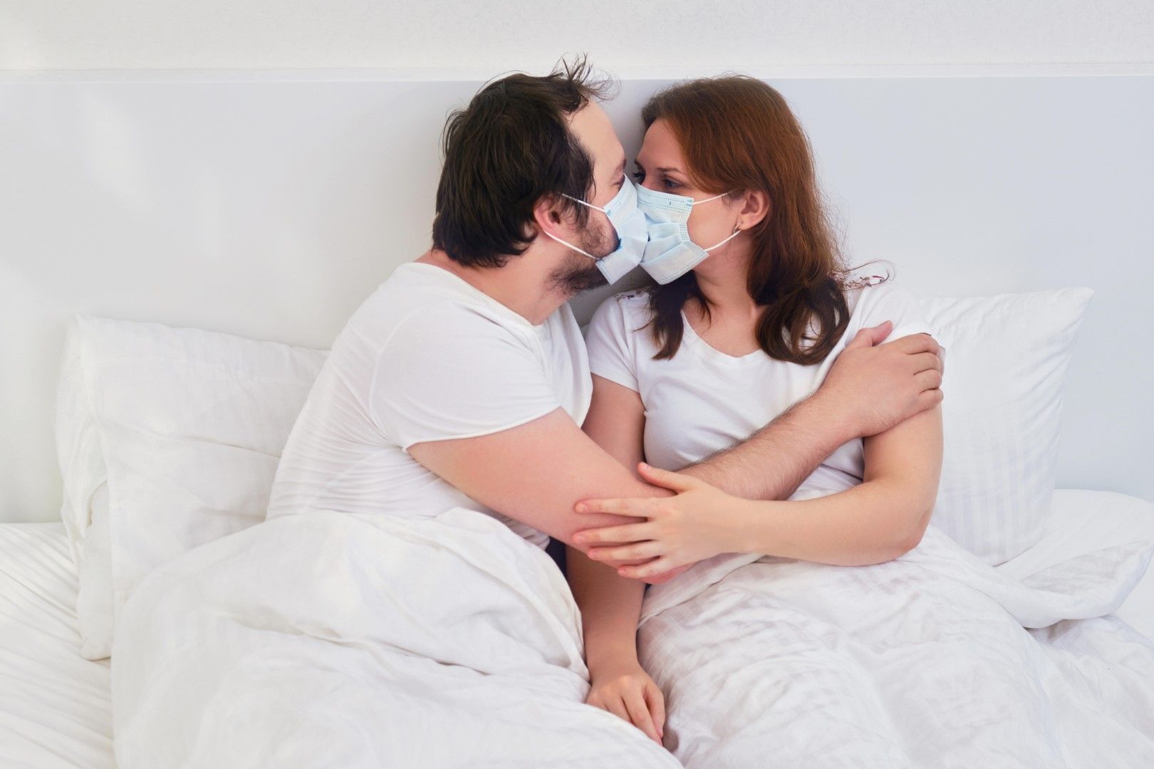 Двойка се целува в леглото с медицински маски. Семейни отношения в изолация заради коронавируса