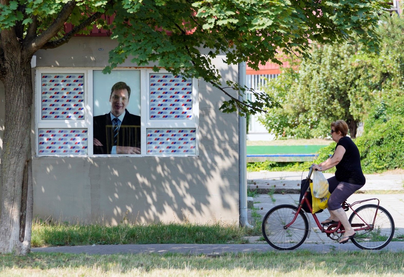 Възрастна жена минава с колео покрай къща, прозорецът на коят е украсен с портрет на президента Вучич