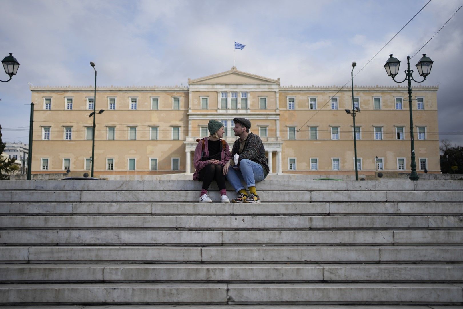 Двойка туристи седят на стълбите на площад Синтагма на фона на парламента в Атина