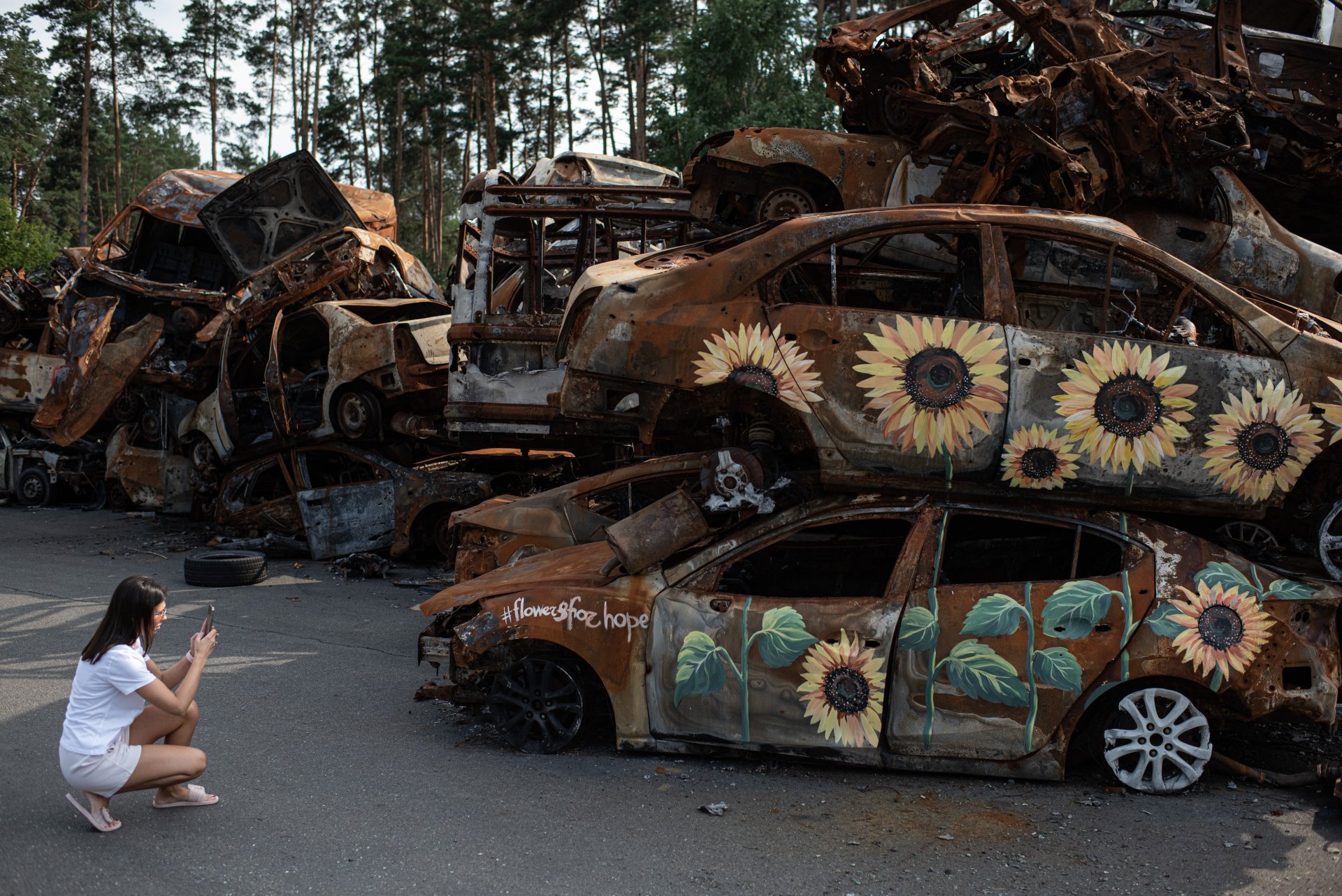 Млада жена прави снимки на изгорели коли, обрисувани със слънчогледи на 17 август 2022 г. в Ирпин, Украйна. 