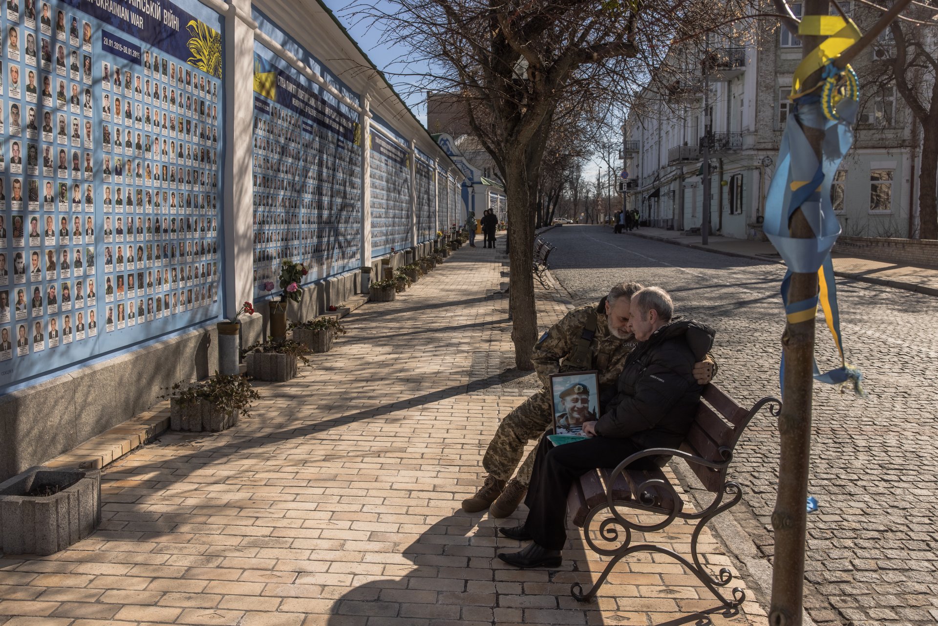 Анатолий седи с портрет на сина си Юрий, украински войник, убит в боевете. Украински боец го прегръща и утешава пред "Стената в памет на падналите защитници на Украйна в Руско- Украинската война". 