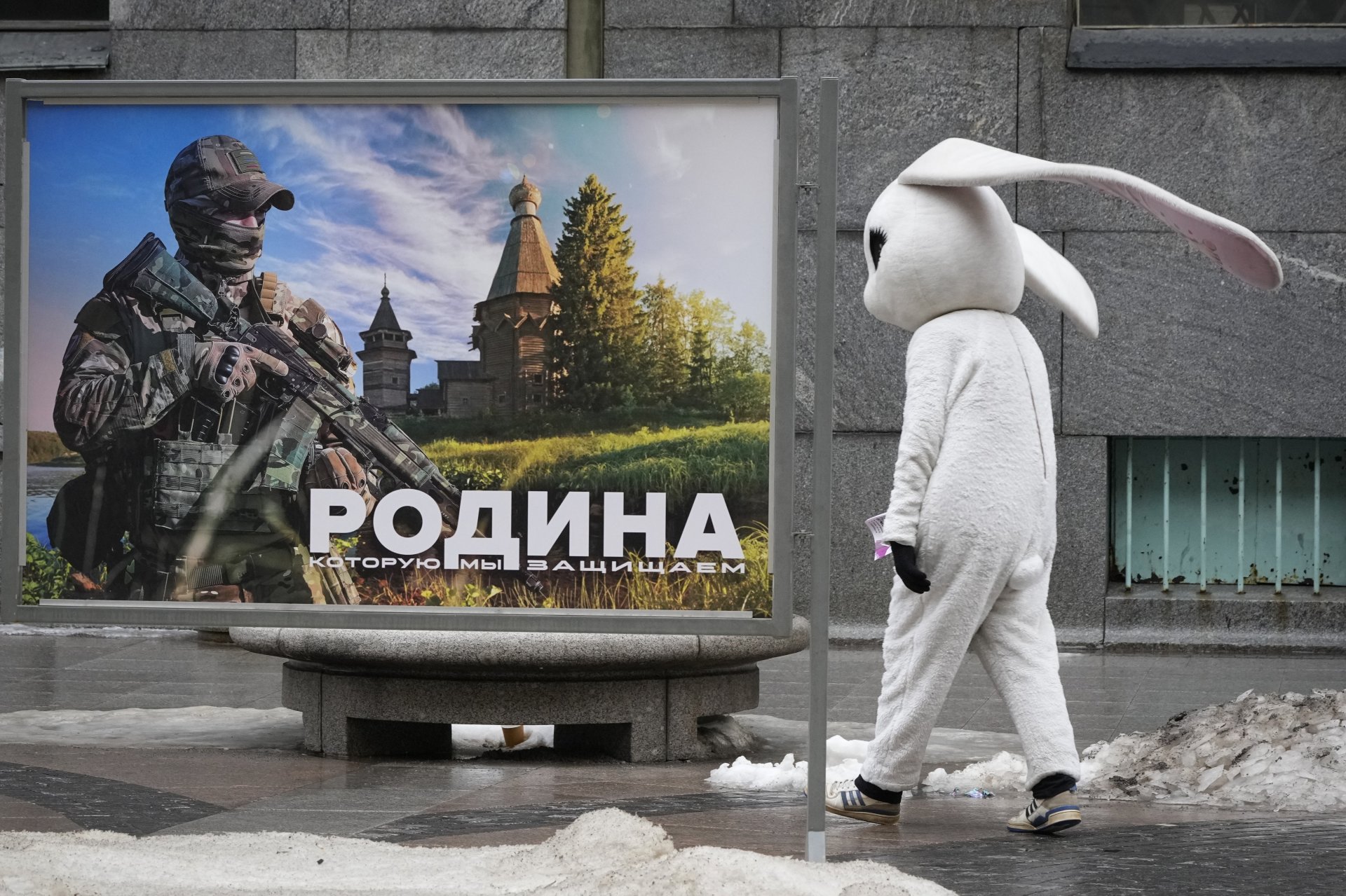 Уличен артист минава покрай плакат на руски военнослужещ, на фона на надпис „Родината, която защитаваме“, на изложба на открито на военни снимки в Санкт Петербург, вторник, 14 март 2023 г.