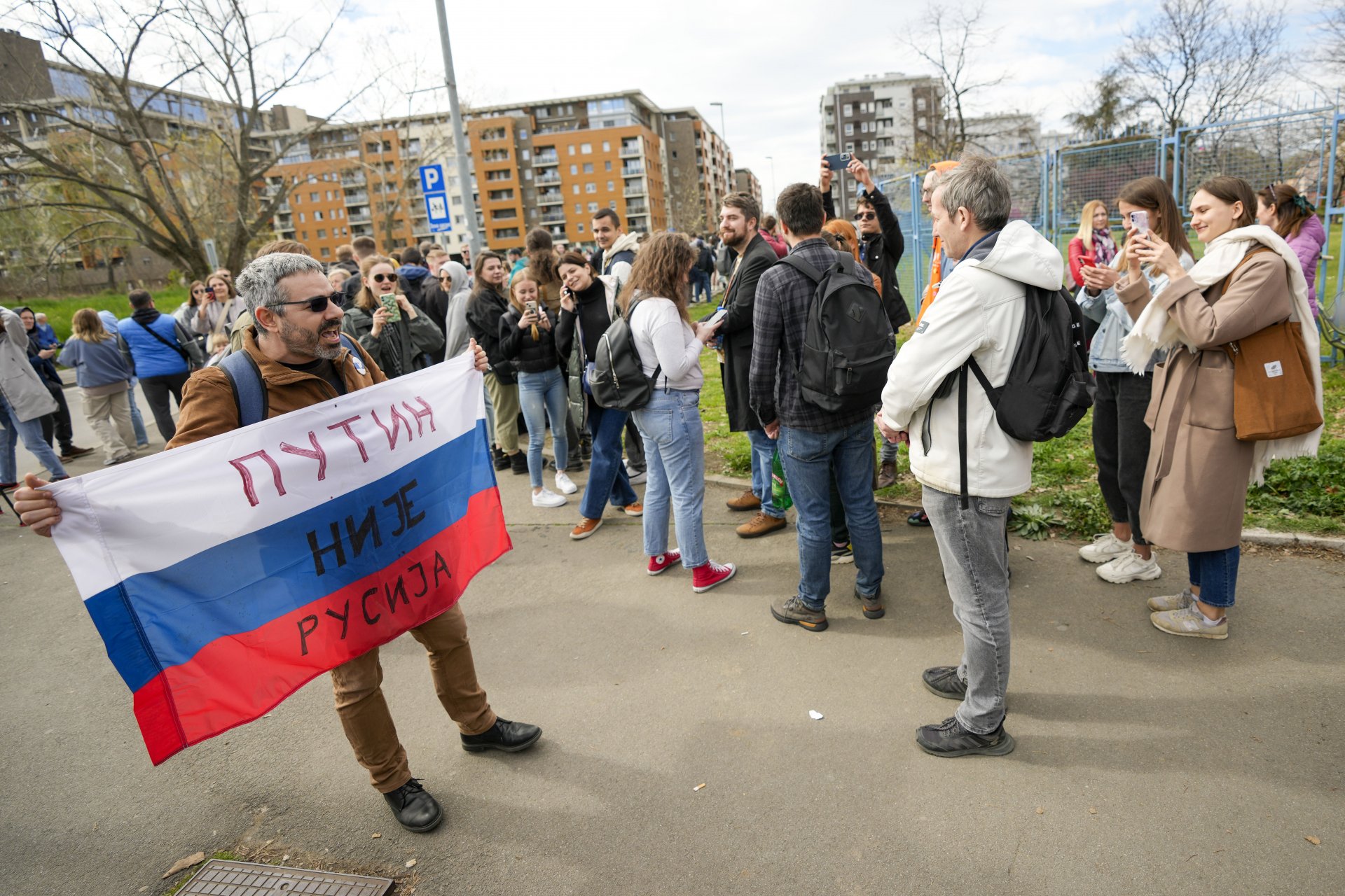 Петър Никитин, руски активист, държи знамето на Русия, на което пише: „Путин не е Русия“ и минава покрай хора, наредили се на опашка, за да гласуват пред избирателна секция, създадена в руско училище в Белград, Сърбия, неделя, 17 март 2024 г.
